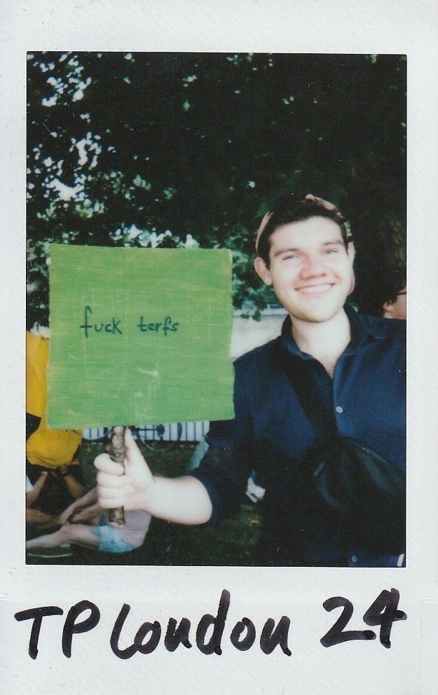 A smiling person is holding a sign with text in a park, standing against a backdrop of trees.