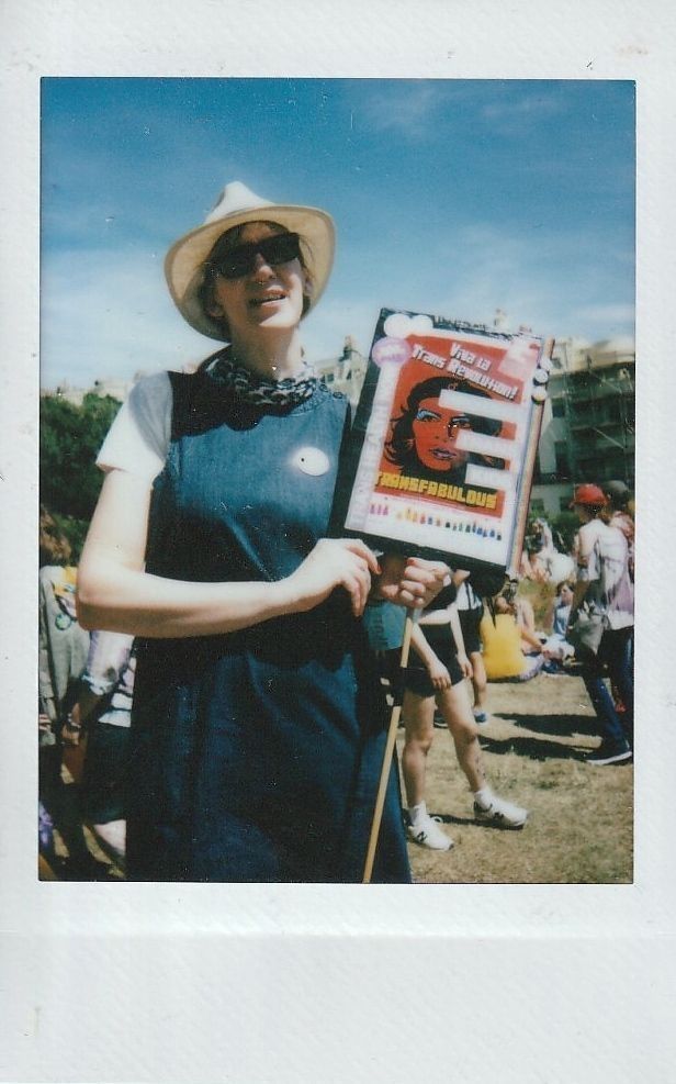 A person in sunglasses and a hat holds a sign which says "Viva La Trans Revolution!".