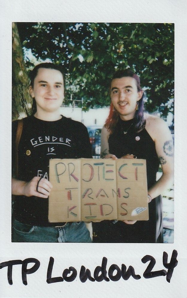 Two people are smiling while holding a cardboard sign that says "Protect Trans Kids" in colorful letters.