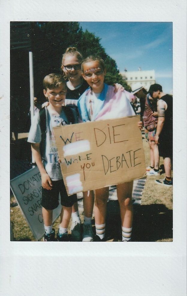 Three people stand together at an outdoor event, holding a sign which says "We Die While You Debate".