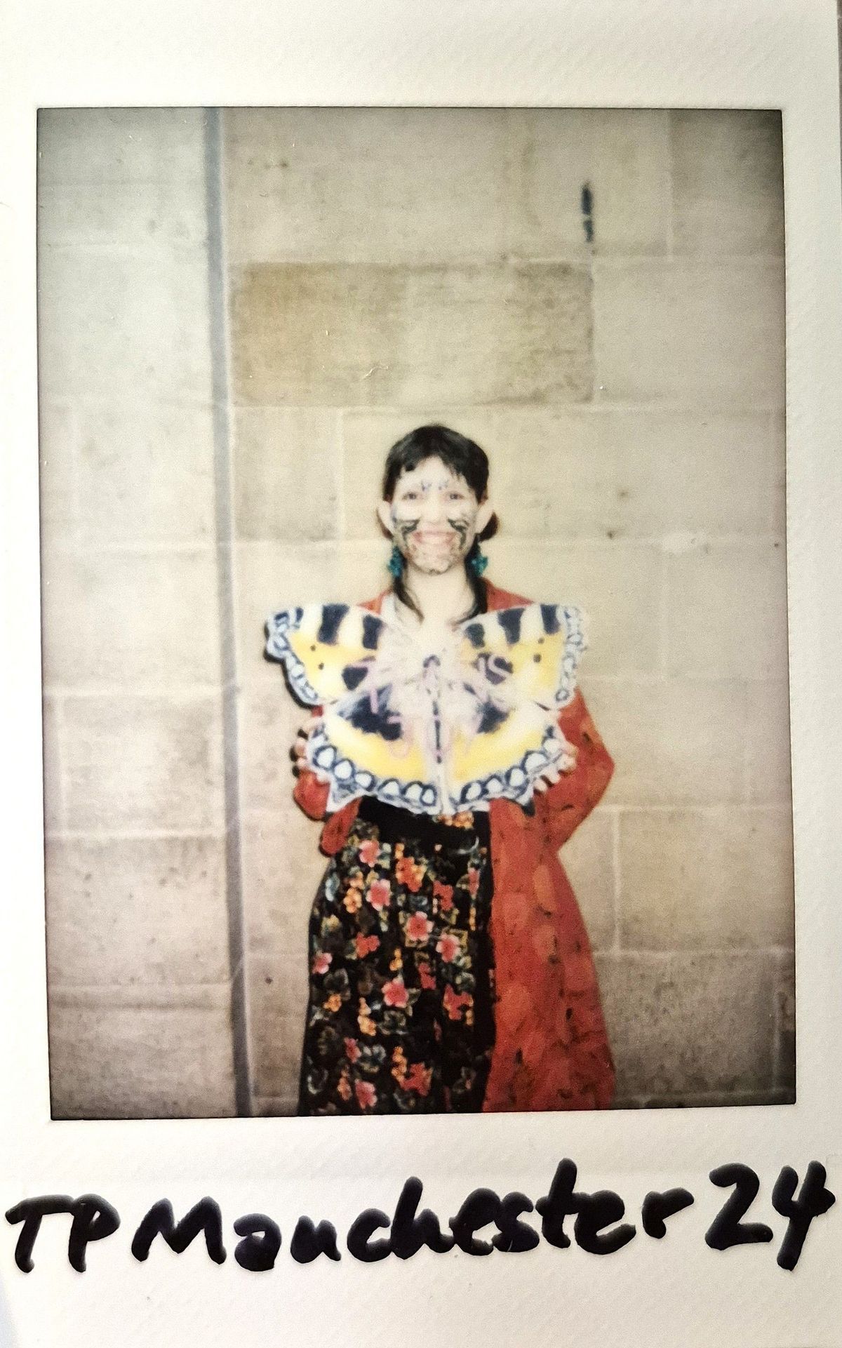 A person stands against a wall, wearing colorful face paint and holding a butterfly-shaped placard.