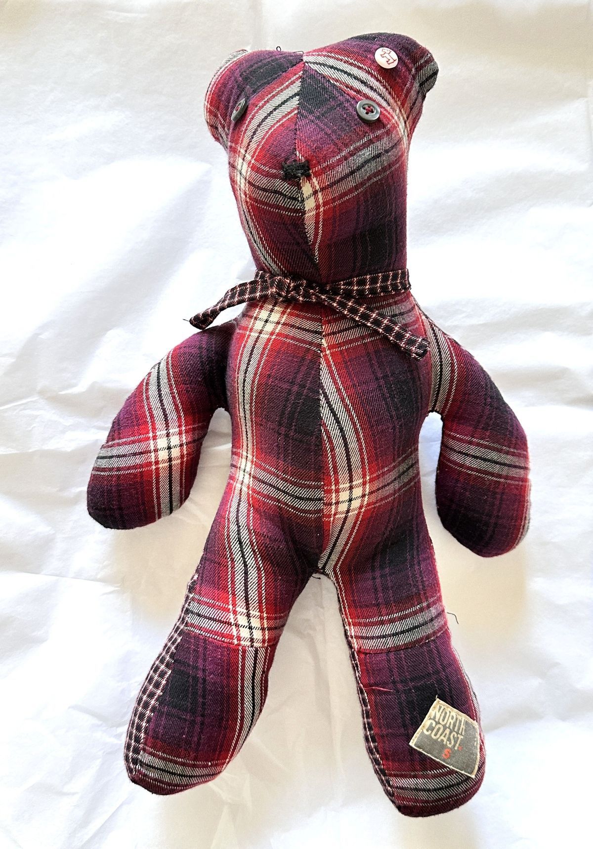 A plaid teddy bear with button eyes and scarf sits on a white background.