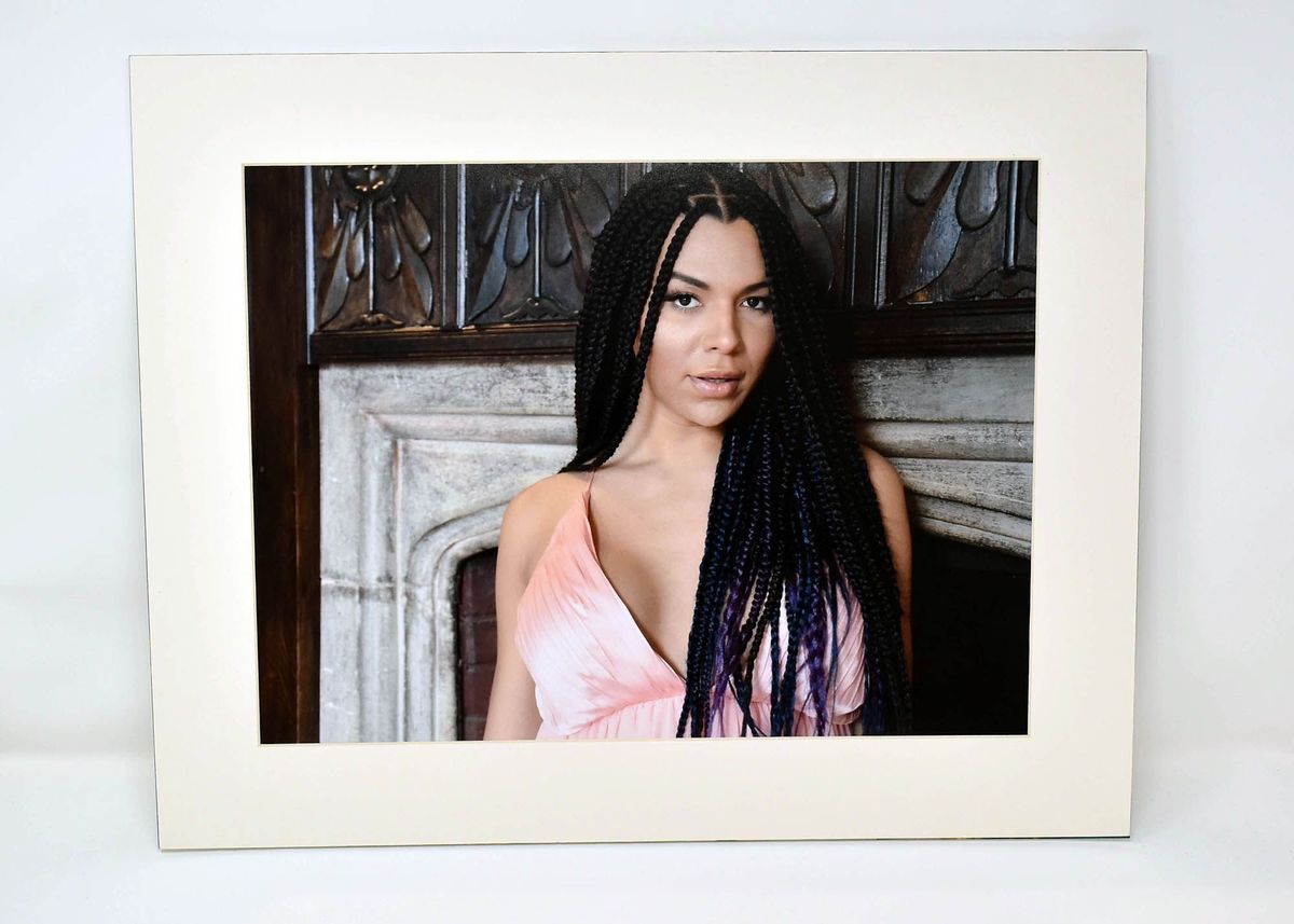 Colour photo of woman with long dark hair braids in pink dress standing in front of fireplace.