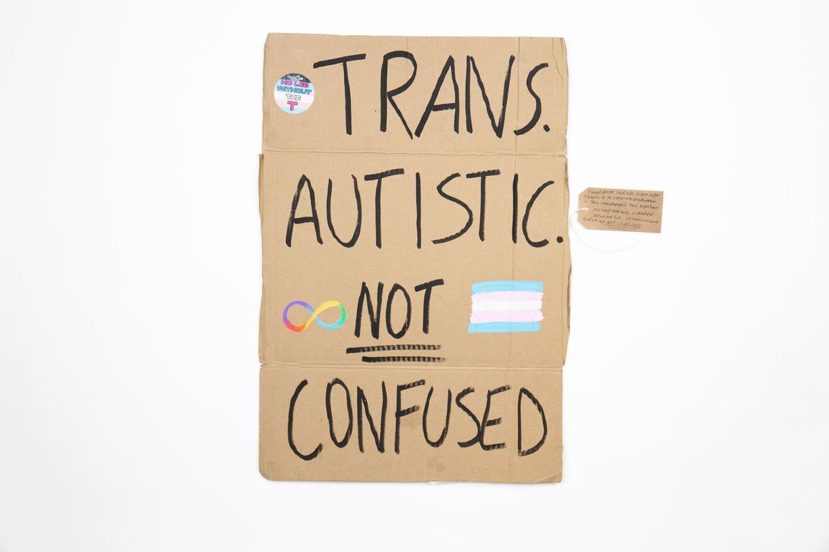 Handwritten cardboard placard reads: "TRANS. AUTISTIC. NOT CONFUSED," ["NOT" is double underlined] with colorful symbols. Decorated with a sticker, a rainbow colored infinity symbol and the trans flag. With a brown, handwritten luggage tag.