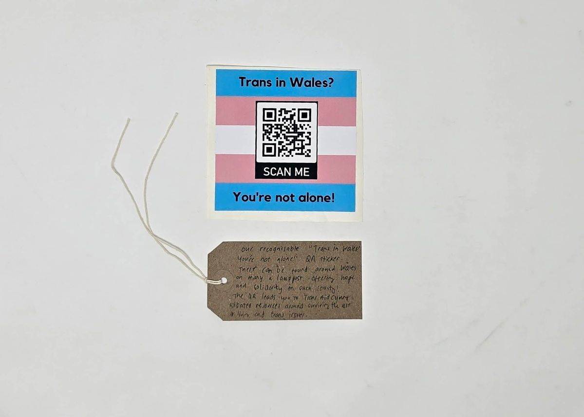 Square sticker in the trans flag colours. There is a QR code in the center and the text "Trans in Wales?" above this and  "You're not alone!" below. Also, a brown hand written luggage tag