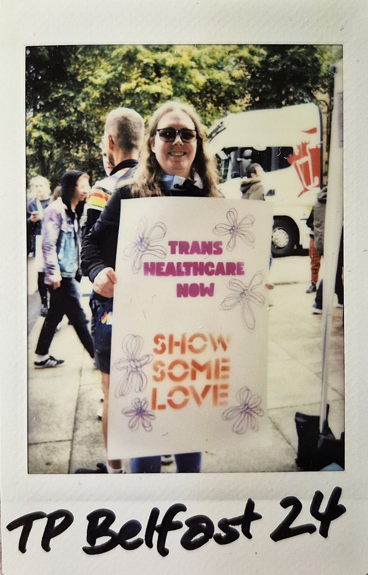 A person smiles while holding a sign advocating for transgender healthcare, surrounded by people on the street.
