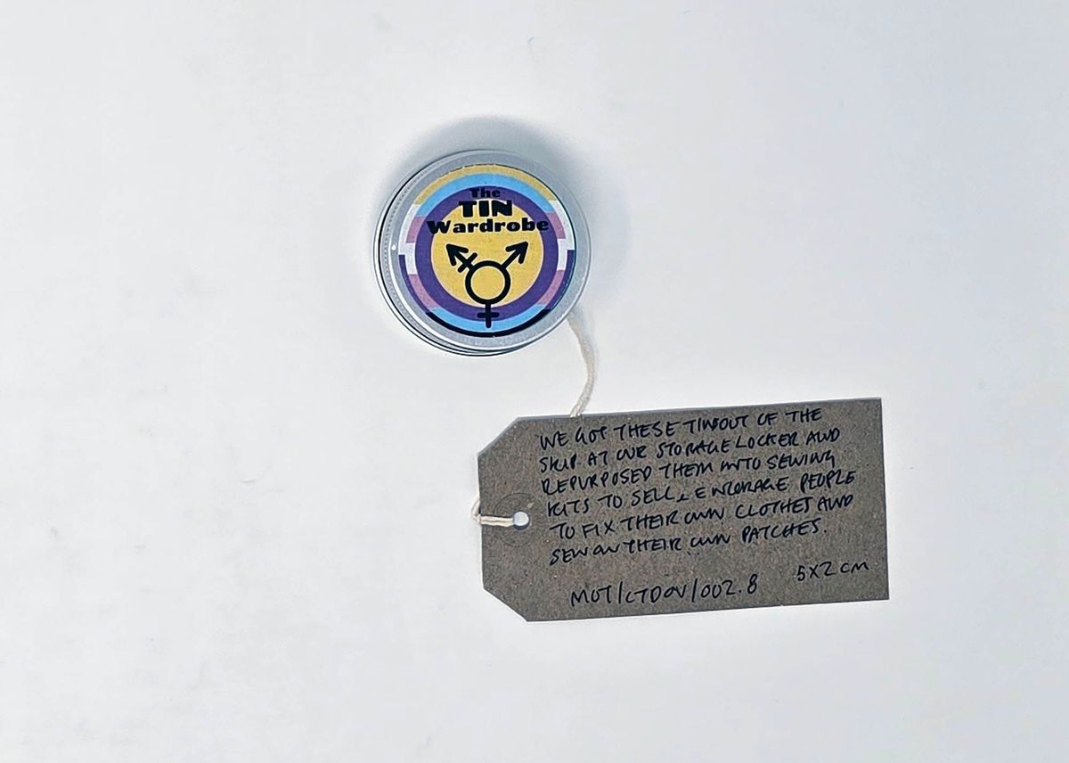 A small, round, silver tin labeled "The TIN Wardrobe". The label is in the trans, non-binary and intersex pride flag colours and features the trans symbol. With a brown handwritten luggage tag.