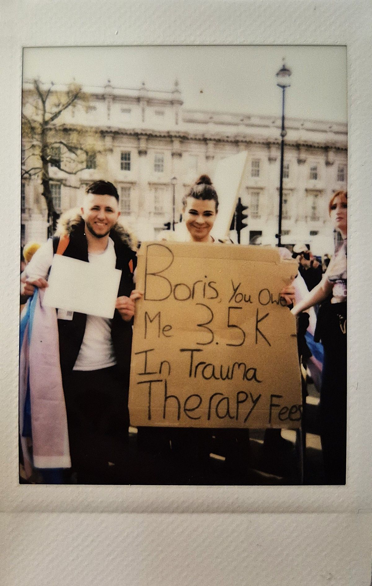 Two people in a protest hold a cardboard sign which says "Boris, you owe me 3.5k in trauma therapy fees".