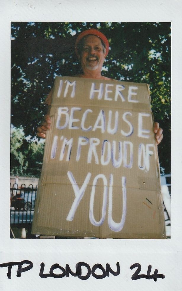 A smiling person holds a supportive sign reading, "I’m here because I’m proud of you,"
