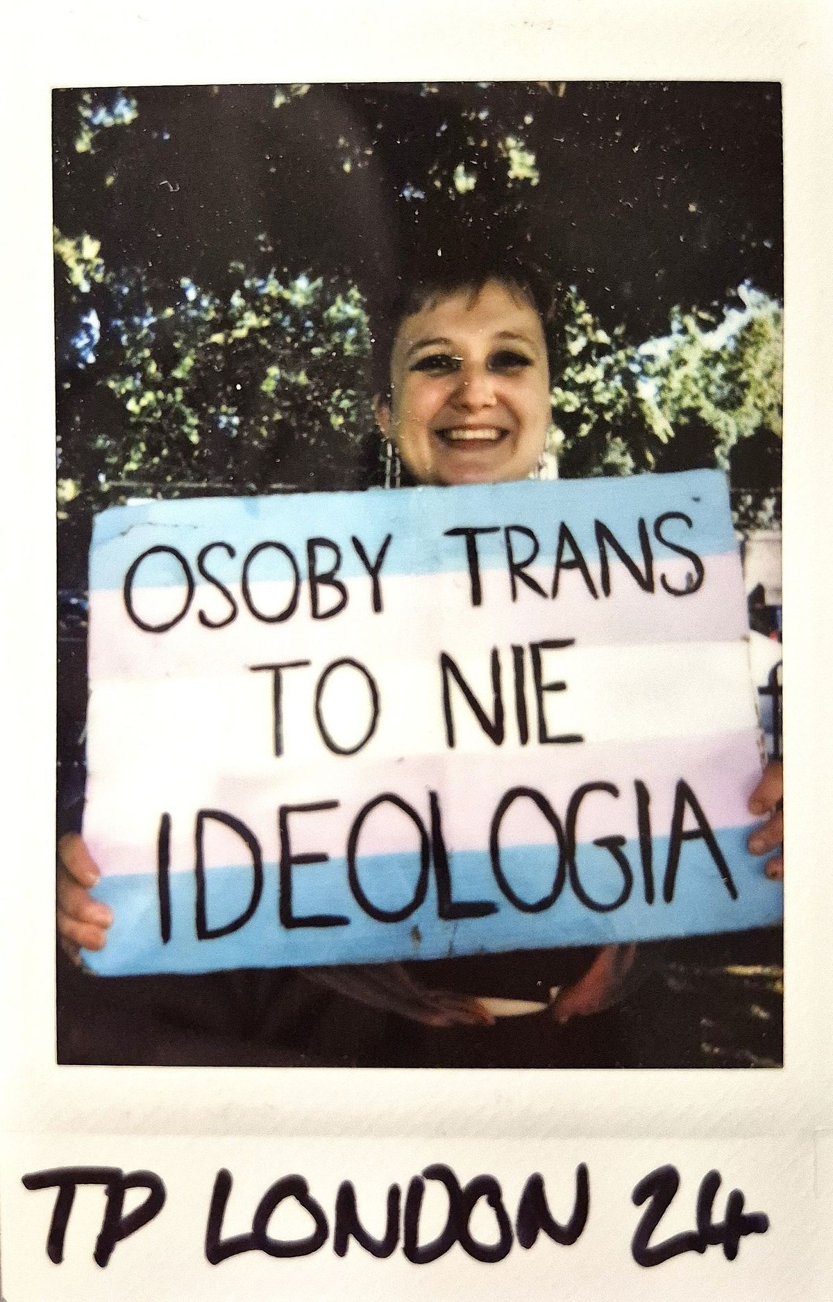 A person holds a sign reading "Osoby Trans to nie ideologia" with a smile outdoors.