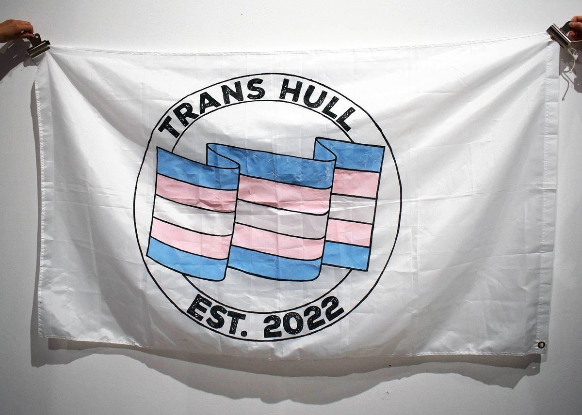 A large white banner with two concentric circles containing the text "TRANS HULL" and "EST. 2022" at the top and bottom. Side the circles is a rippling blue, white and pink trans pride flag