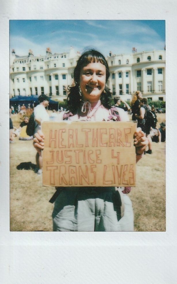 A person smiles holding a sign that reads "Healthcare Justice 4 Trans Lives" at an outdoor gathering.
