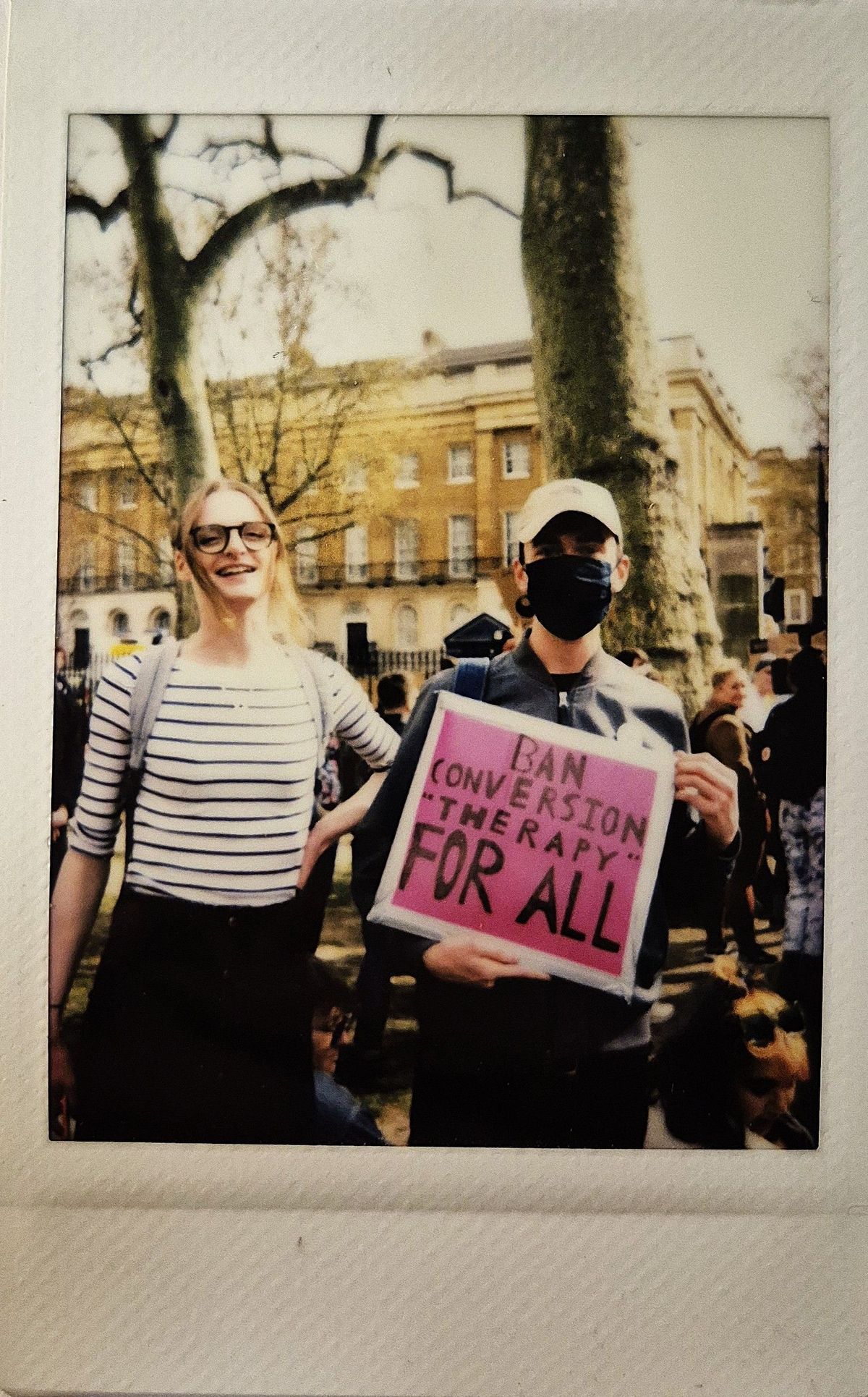 Two people are standing outdoors, holding a sign that says, "Ban Conversion Therapy for All."