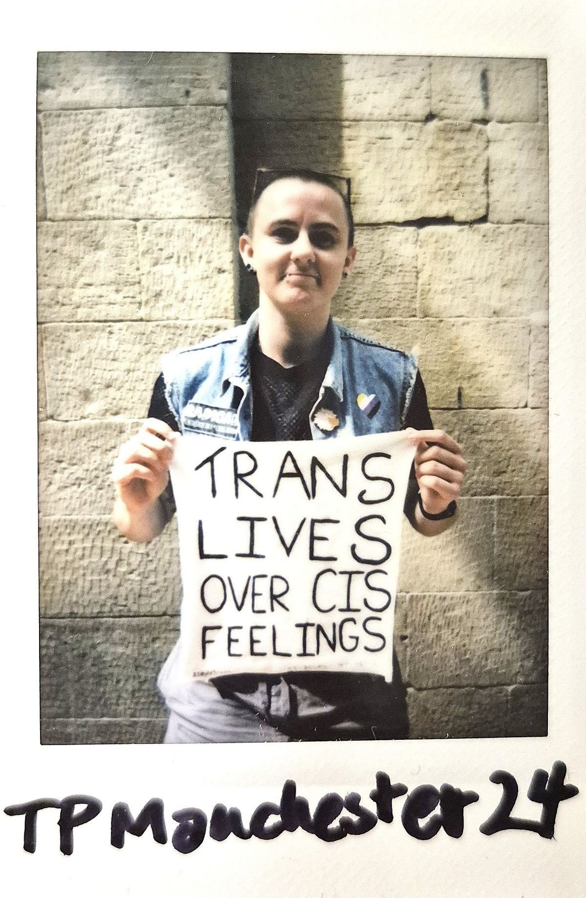 A person stands against a stone wall, holding a sign that reads, "TRANS LIVES OVER CIS FEELINGS".