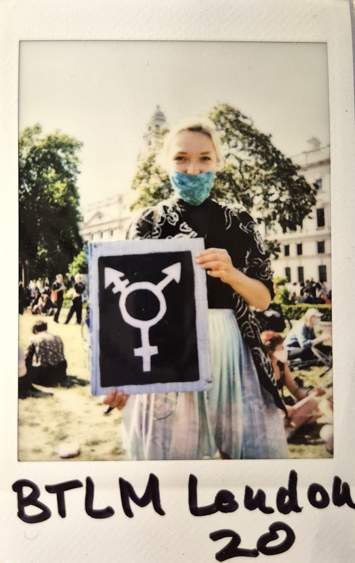 A person wearing a mask holds a sign with a gender symbol at an outdoor event.