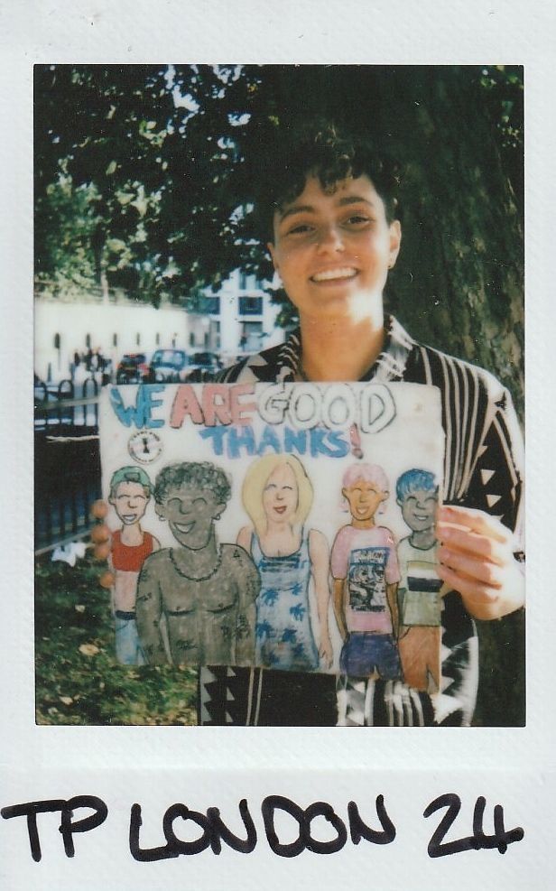 A person smiles while holding a colorful drawing reading, "We are good thanks!" in an outdoor setting
