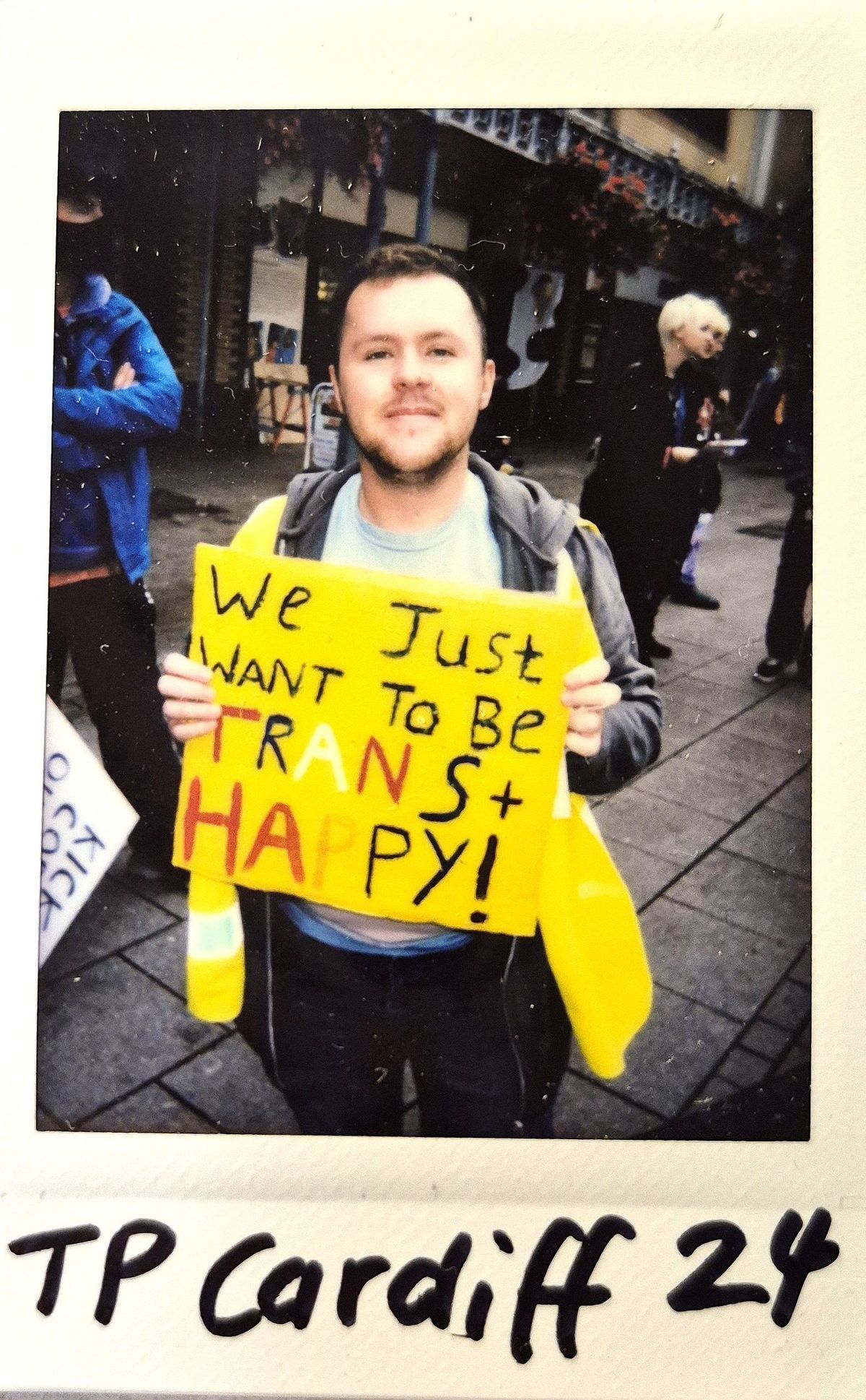 A person holds a bright yellow sign reading "We just want to be trans + happy!"