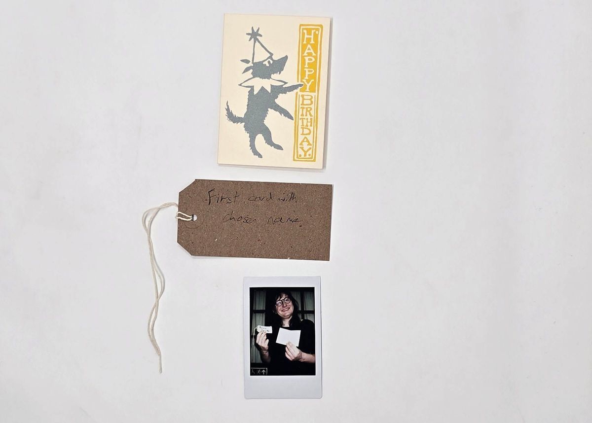 A yellow birthday card depicting a grey dog wearing a ruff and a pointy hat. With a brown, handwritten luggage tag and a polaroid of a person holding the card.