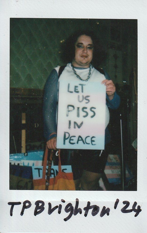 A person is holding a sign advocating for bathroom rights, stating, "LET US PISS IN PEACE".