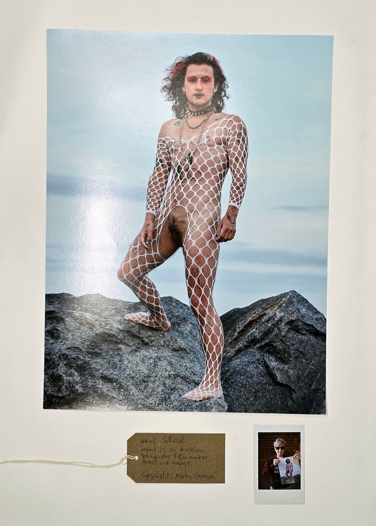 A person who is wearing a white fishnet bodysuit and is stood on rocks in front of the ocean.