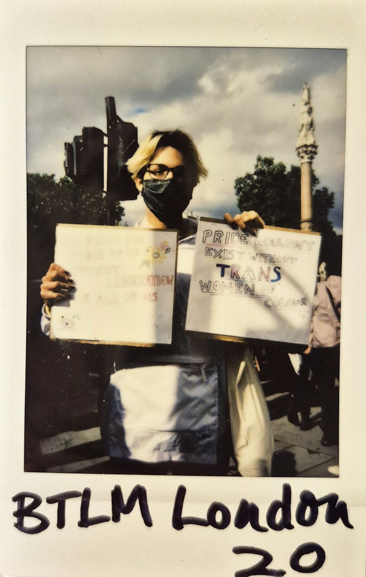 A person in a mask holds two protest signs supporting trans rights at a London event.