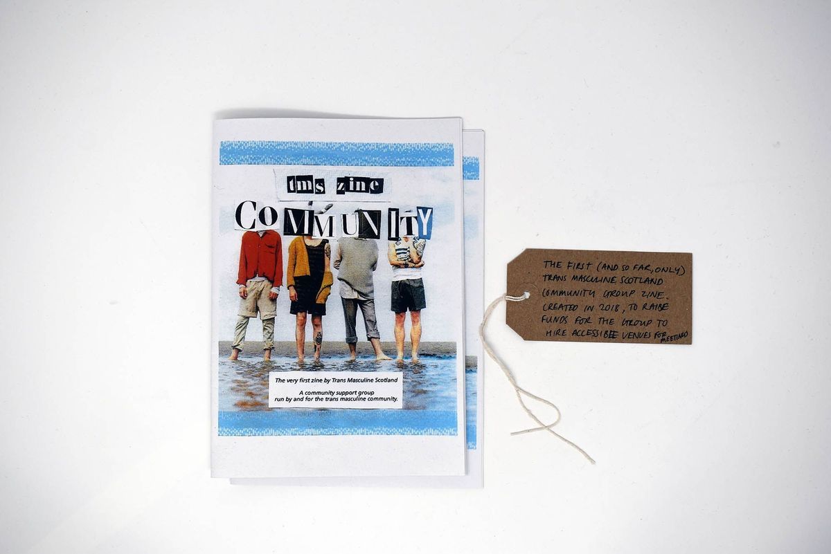 Two copies of the Trans Masculine Scotland (TMS) Community Group zine. Cover image is 4 people paddling in water with the title obscuring their faces. With a brown handwritten luggage tag.