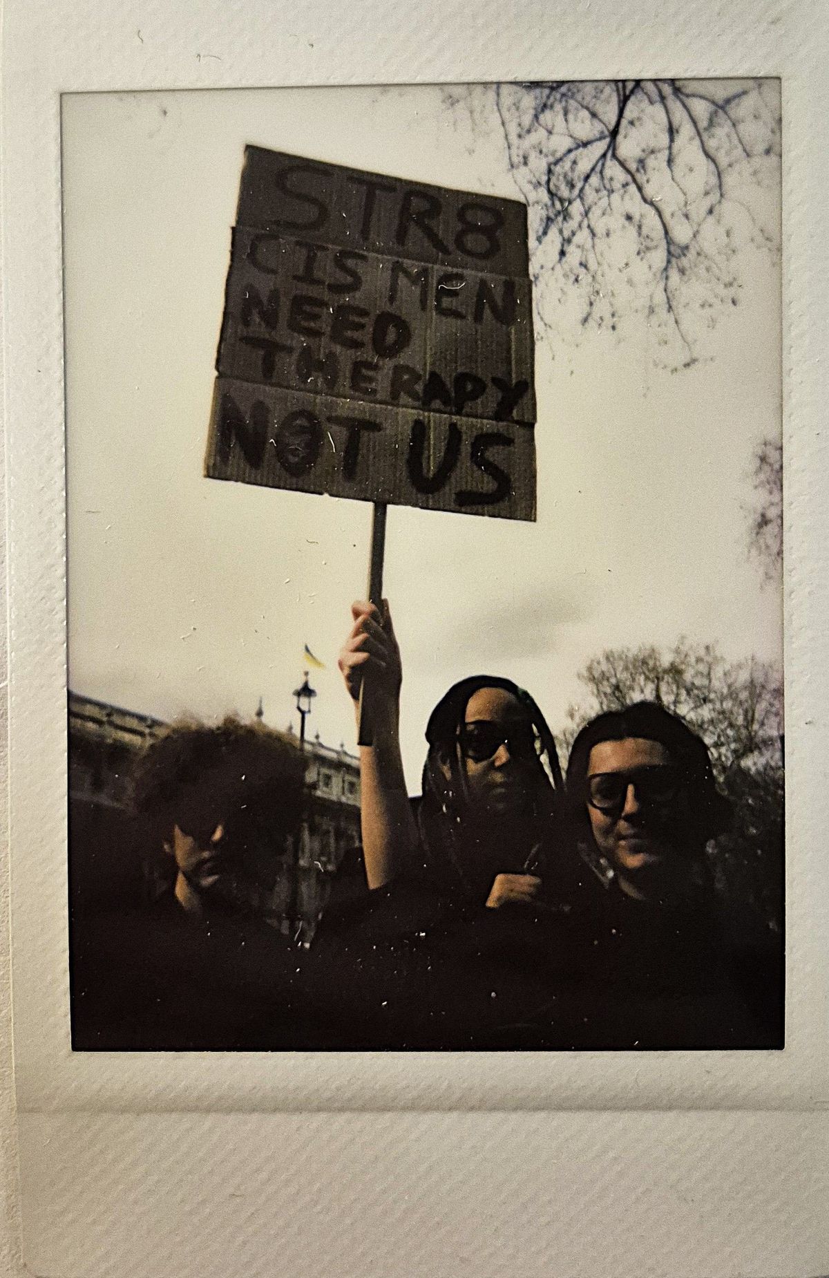 Three individuals stand together, holding a protest sign that reads, "STR8 CIS MEN NEED THERAPY NOT US".