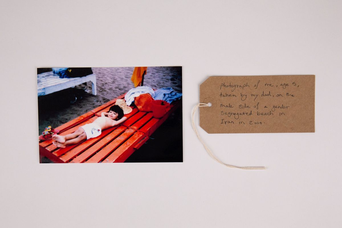 A young, dark haired child wearing white shorts lies on a red, slatted, wooden sunbed on a sandy beach. Their legs are crossed at the ankles and their hands are behind their head. With a brown, handwritten luggage tag.