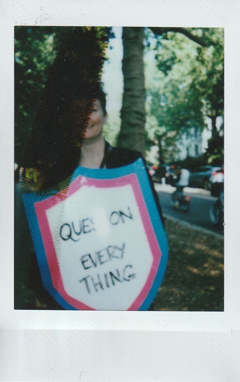 A person partially obscured by a shadow holds a shield-shaped sign saying "QUESTION EVERYTHING" outdoors.