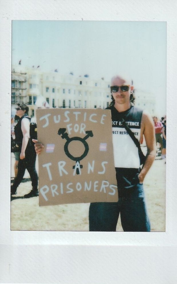 A person is holding a sign reading "Justice for Trans Prisoners" at an outdoor event.