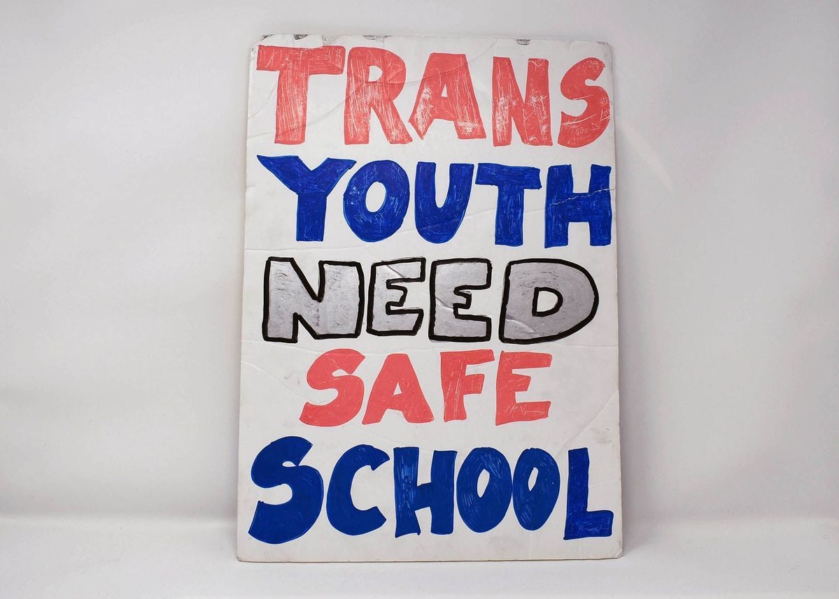 Hand painted, white, cardboard placard reads: "TRANS YOUTH NEED SAGE SCHOOL" in red, blue and silver