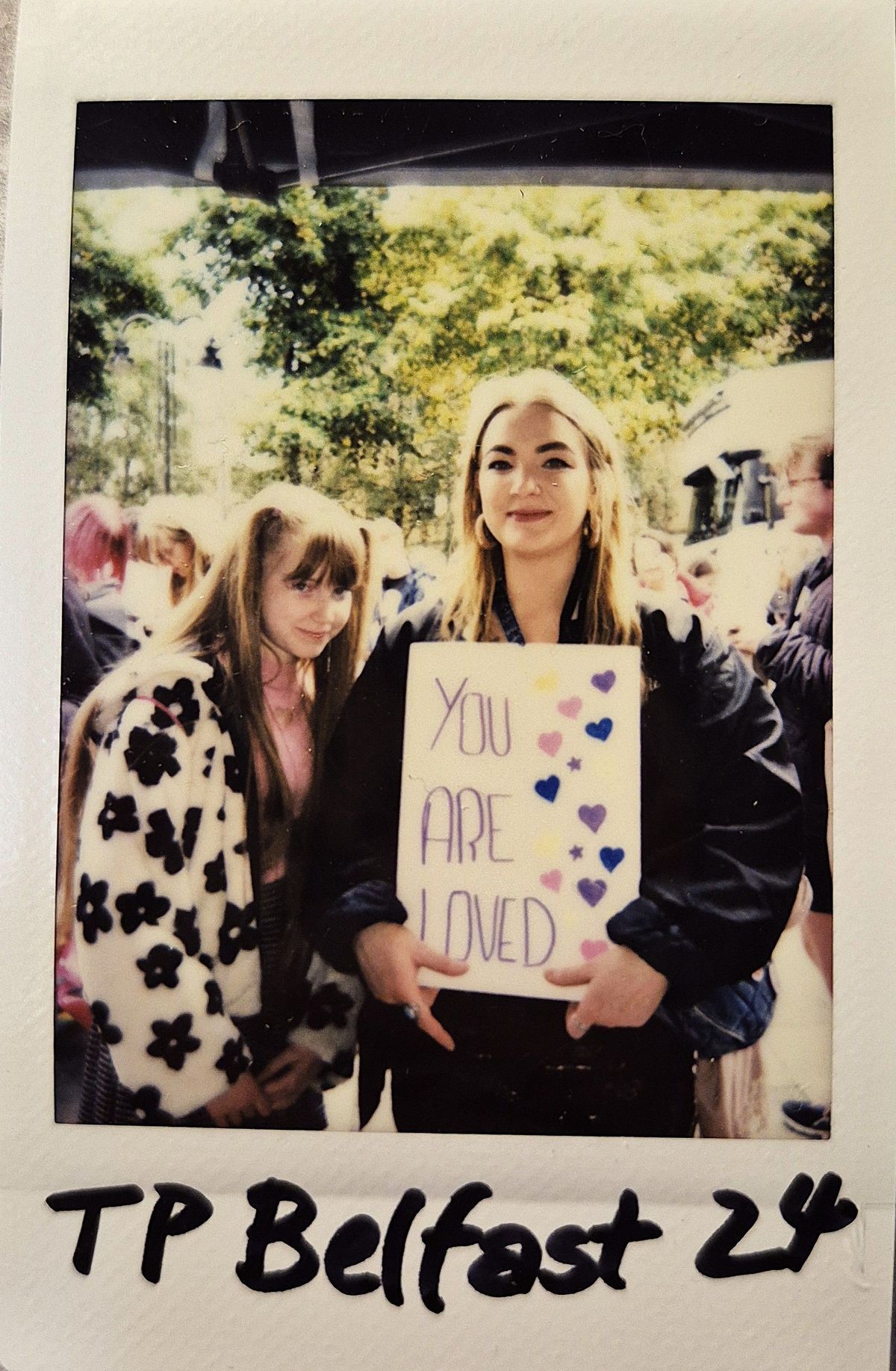 Two people stand outside, with one holding a sign that says, "You Are Loved."