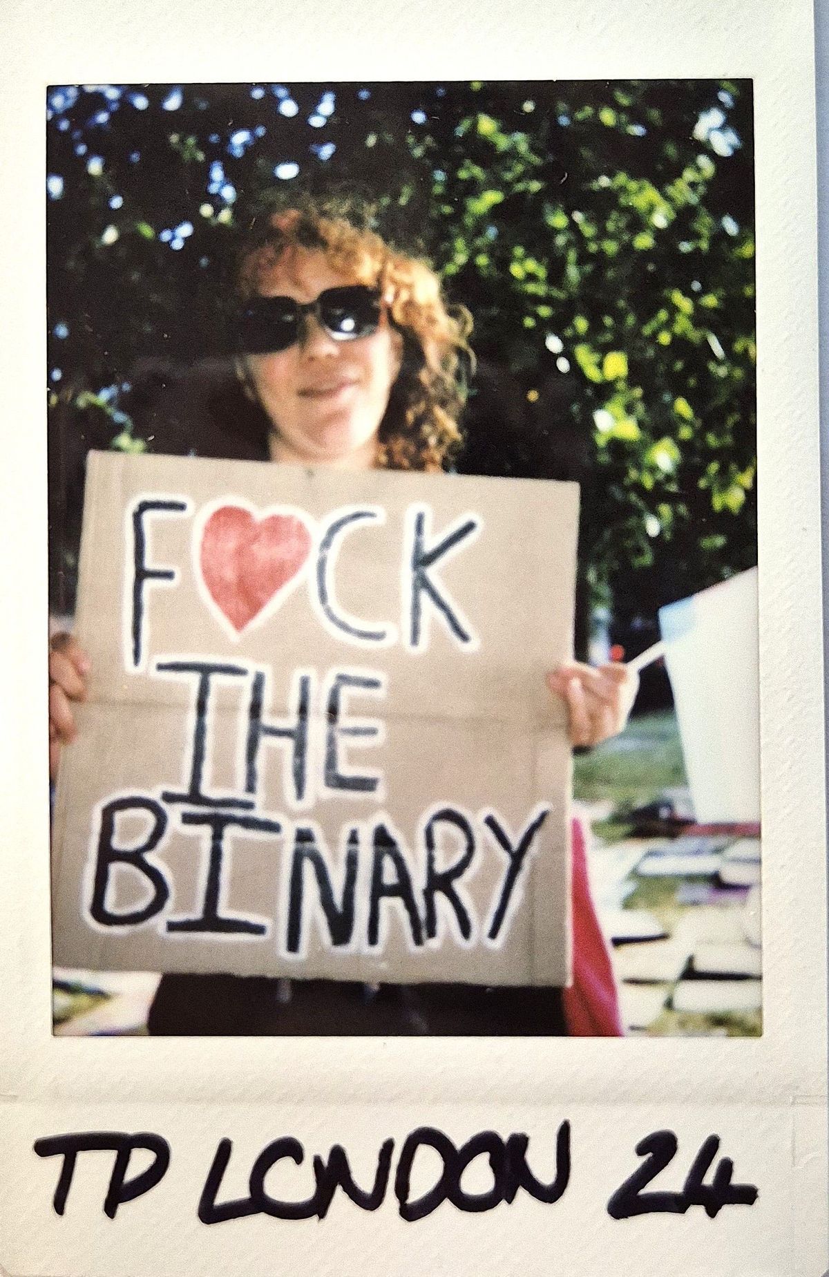 A person wearing sunglasses holds a sign saying "F♥CK THE BINARY" at an outdoor event