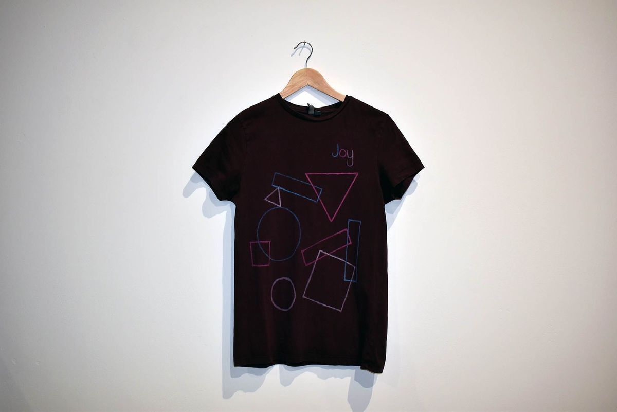 A burgundy t-shirt, printed with colored line drawings of geometric shapes and the word "Joy", on a wooden hangar.