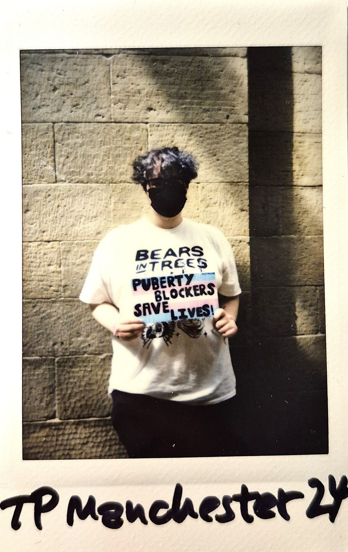 A person wearing a mask stands by a stone wall with a placard.