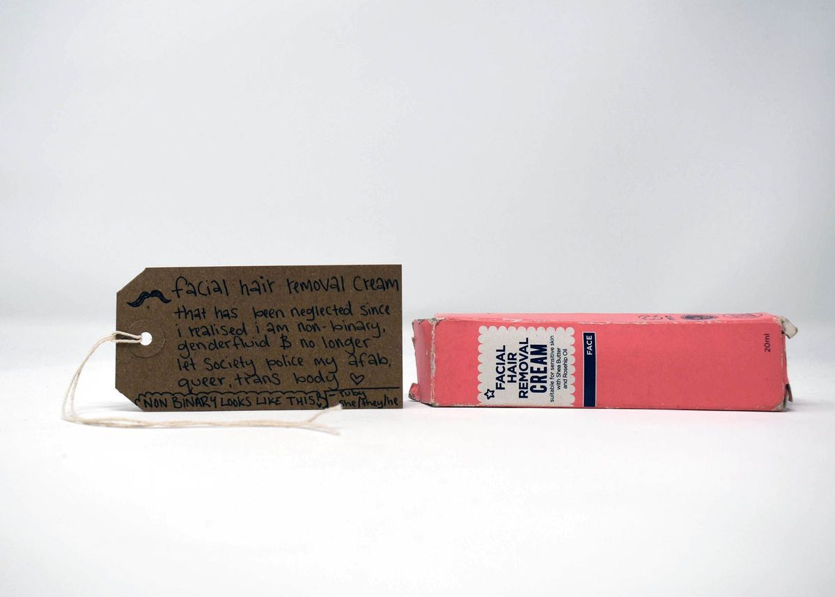 A facial hair removal cream box sits beside a tag expressing non-binary and genderfluid identities.