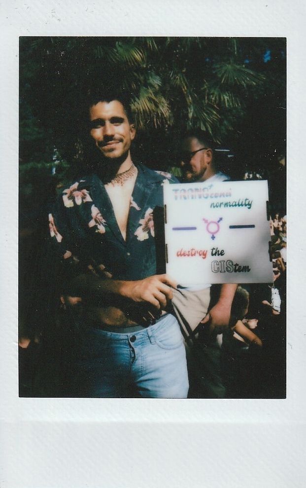 A smiling person in a floral shirt holds a sign which says something that is slightly blurry and hard to make out.