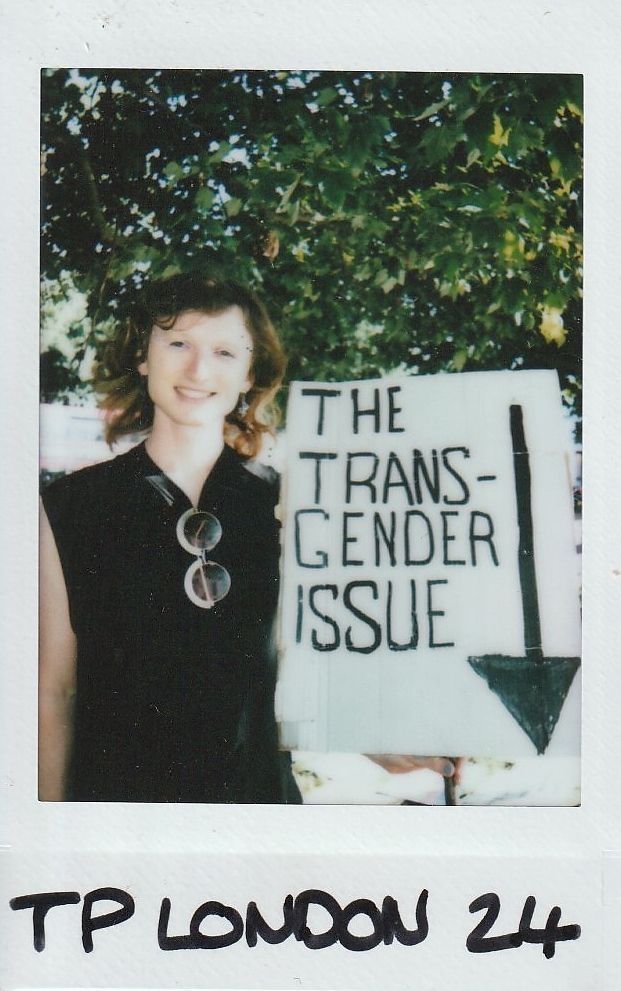 A person is standing outdoors, smiling beside a sign reading "THE TRANSGENDER ISSUE".