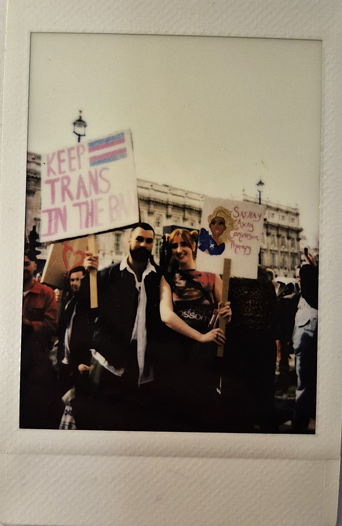 Two people hold signs which say "KEEP TRANS IN THE BAN" and "SASHAY AWAY CONVERSION THERAPY".