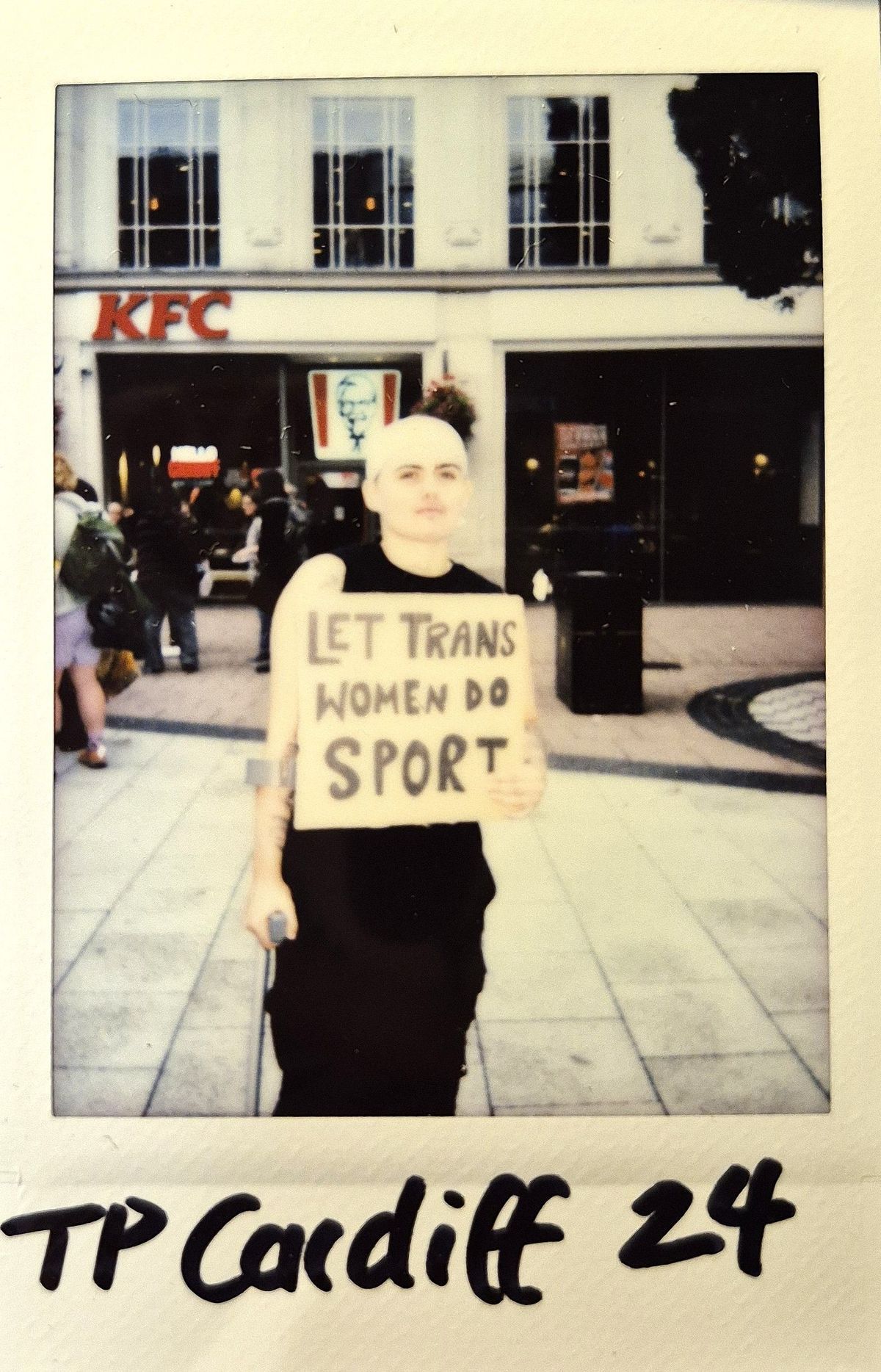 A person stands in front of a building, holding a sign supporting trans women's participation in sports.
