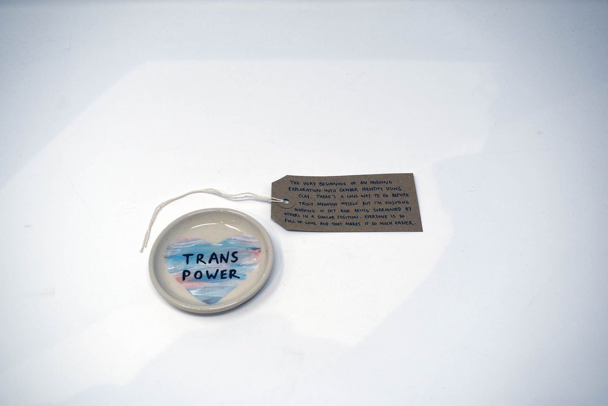 A small, white, ceramic dish illustrated with a blue, pink and white heart and the text "TRANS POWER" in black. With a brown, handwritten luggage tag.
