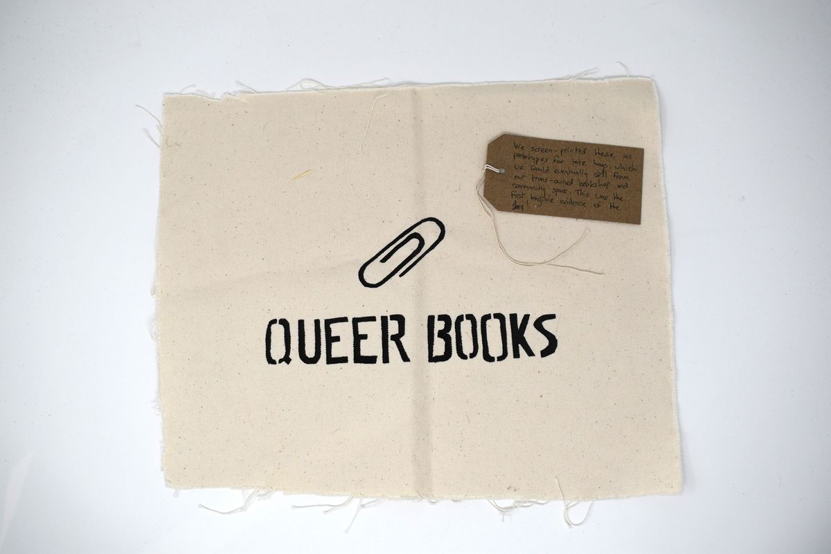 A square piece of fabric displays the words "Queer Books" with a paperclip icon above.