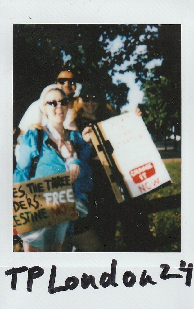 Three people are holding signs with messages during a protest event in a park setting.