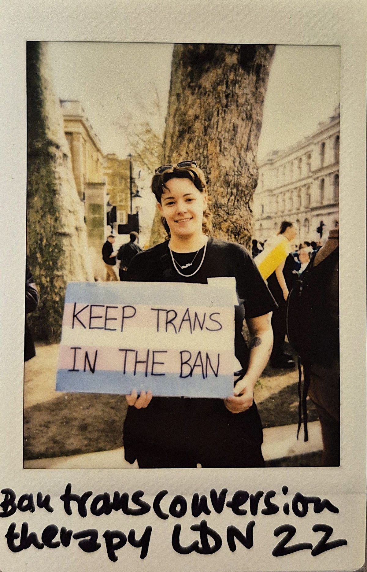 A person holding a sign saying “KEEP TRANS IN THE BAN” is standing outdoors, smiling.