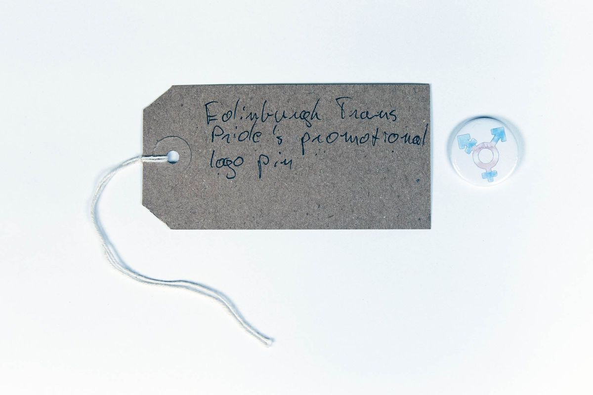 A small, round badge featuring the trans symbol in the trans colours on a white background. With a brown, handwritten luggage tag.