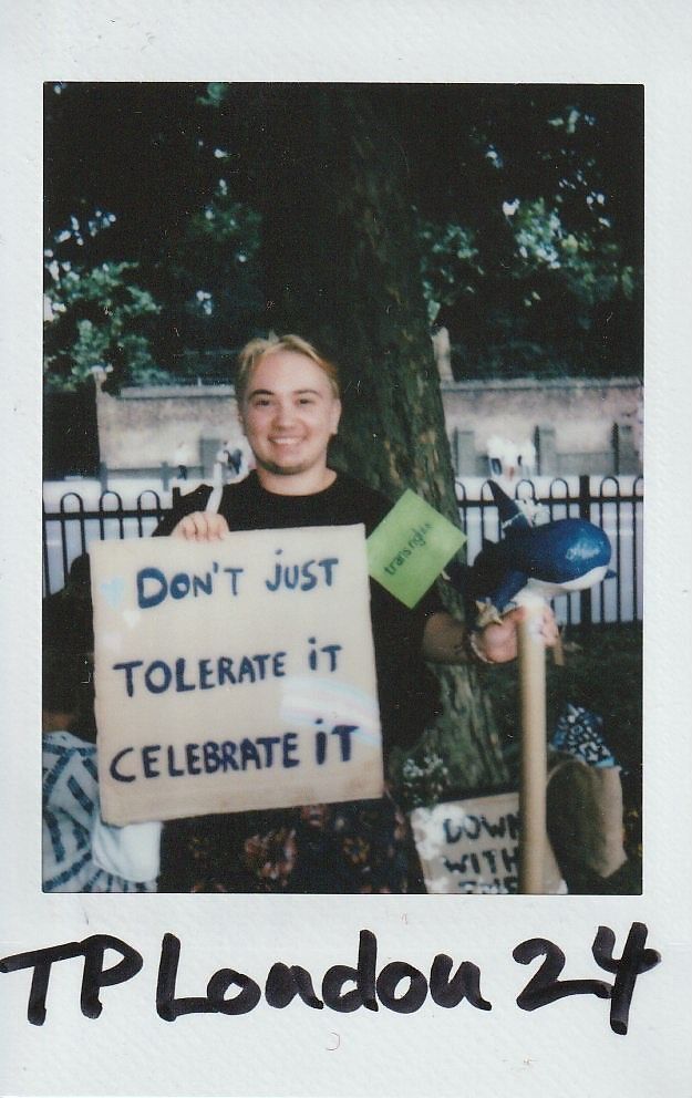A person stands smiling, holding a sign that reads, "Don't just tolerate it, celebrate it".