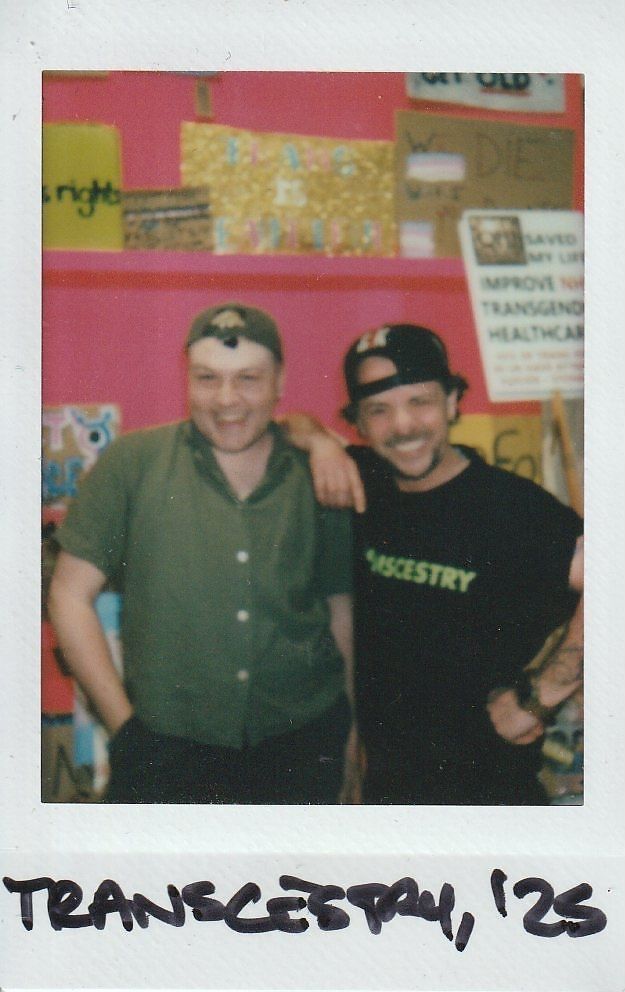 Two people stand smiling in front of a vibrant pink background with various posters.