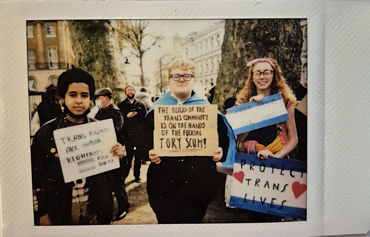 Three people are holding signs advocating for trans rights at a public protest. One says "TRANS RIGHTS ARE HUMAN RIGHTS FUCK BOJO", one says "THE BLOOD OF THE TRANS COMMUNITY IS ON THE HANDS OF THE FUCKING TORY SCUM" and ones says "PROTECT TRANS LIVES".