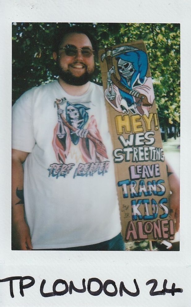 A person wearing sunglasses holds a sign reading "Leave Trans Kids Alone!" in a park setting.