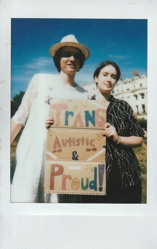 Two people stand together outdoors, holding a colorful sign reading "Trans Autistic & Proud".